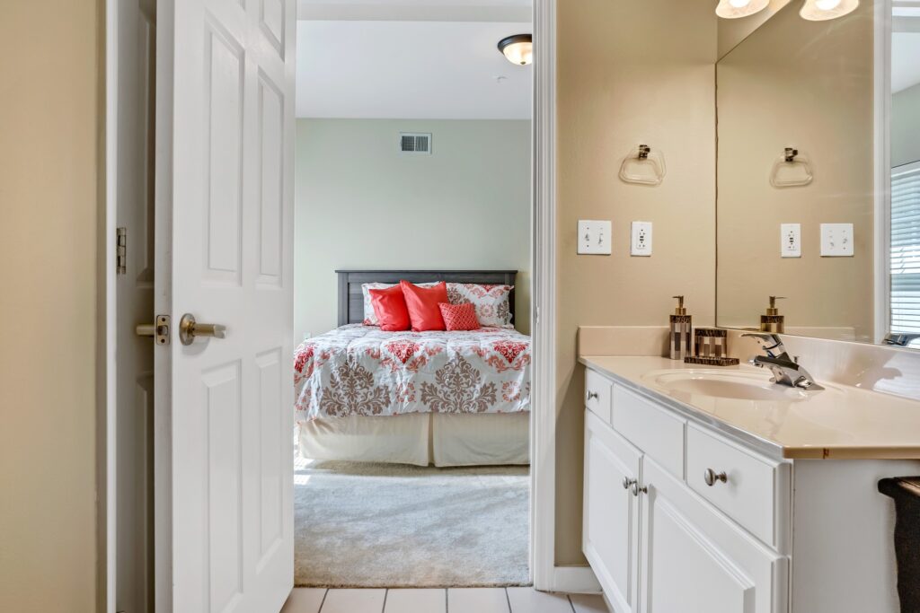 AL101282_2385_Dolly_Ridge_Vestavia_20220531_069 - Town Village Vestavia Hills A bathroom with a sink and white counters and an open door that opens the view into the bedroom with a made-up bed with red and white sheets and red throw pillows at Town Village Vestavia Hills, a senior living community near Homewood, AL.
