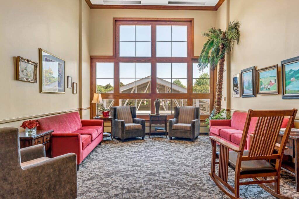 A sitting area with two red couches facing one another, two sofa chairs against a ceiling-high window with a side table between them and a lamp on it, a brown sofa chair, and a rocking chair at Town Village Vestavia Hills, a senior living community.