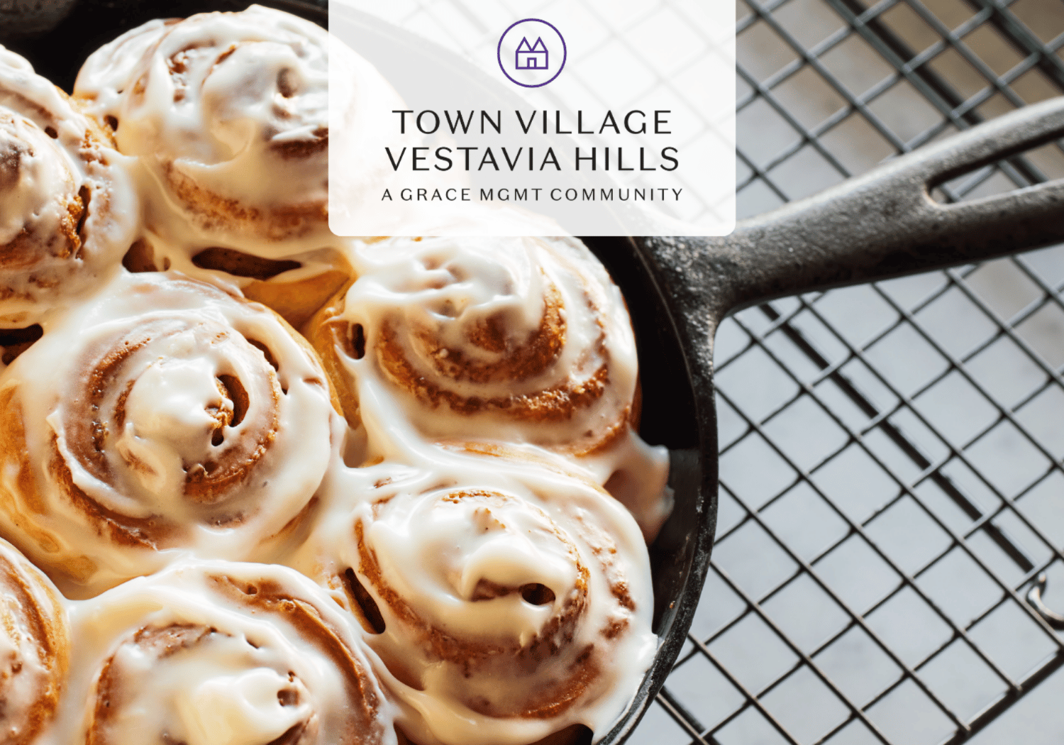 Cast iron skillet filled with glazed cinnamon rolls, with a translucent overlay showing the Town Village Vestavia Hills logo and text above the rolls.